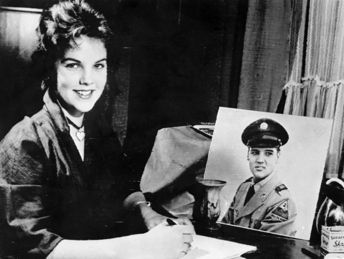 A black and white picture of Priscilla Presley writing a letter while sitting next to a photo of Elvis in his Army uniform.