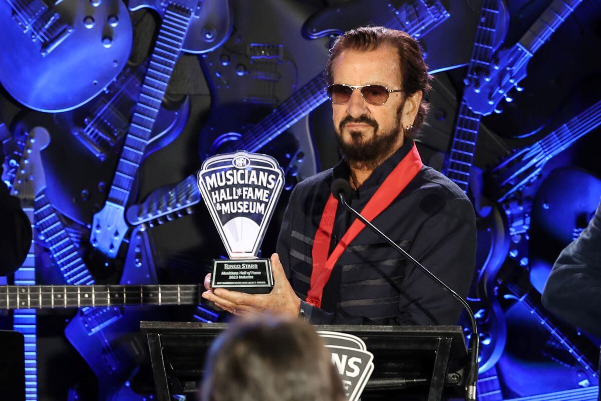 Ringo Starr wears a red ribbon around his neck and holds a "Musician's Hall of Fame & Museum" statue. He stands at a podium.