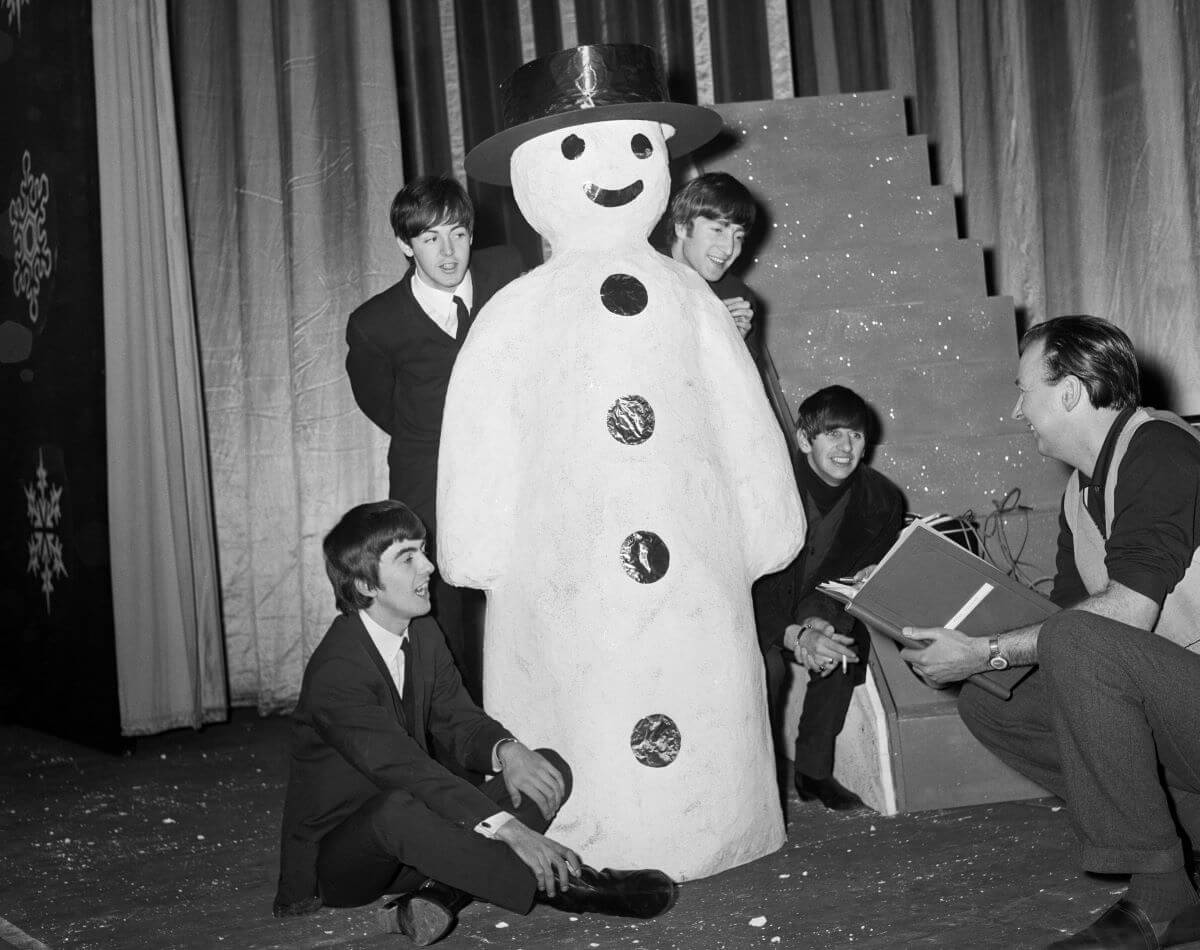 A black and white picture of The Beatles wearing suits and gathering around a snowman statue. They stand on a stage.