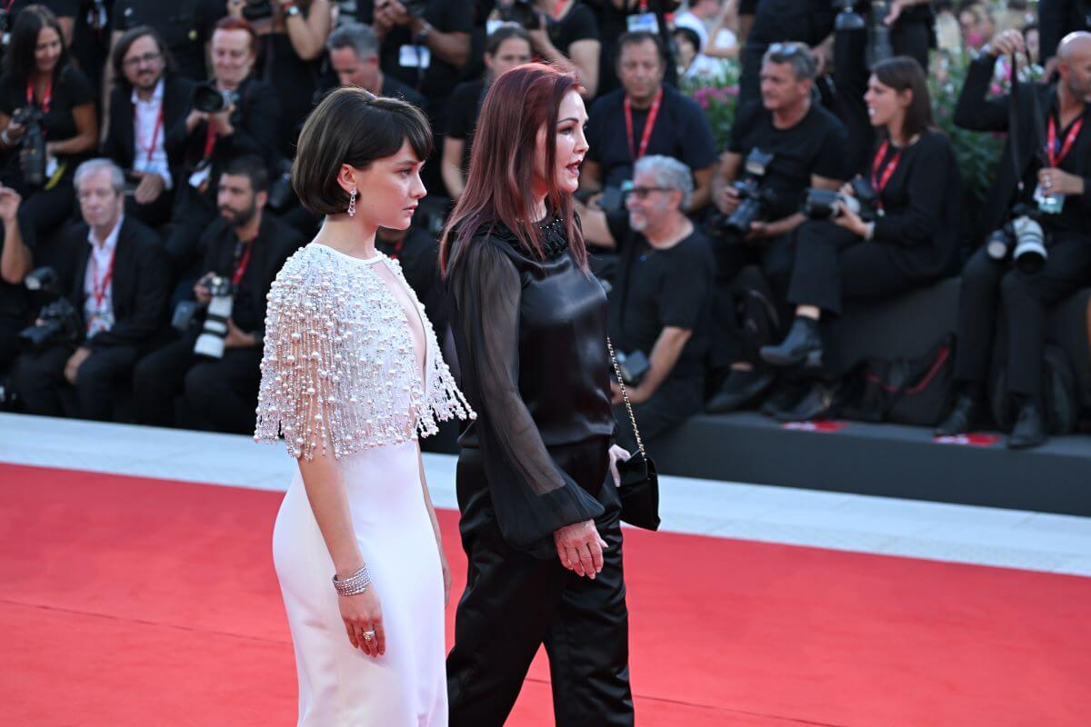 Cailee Spaeny and Priscilla Presley walk down a red carpet together. Spaeny wears a white dress and Priscilla wears black pants and a black shirt.