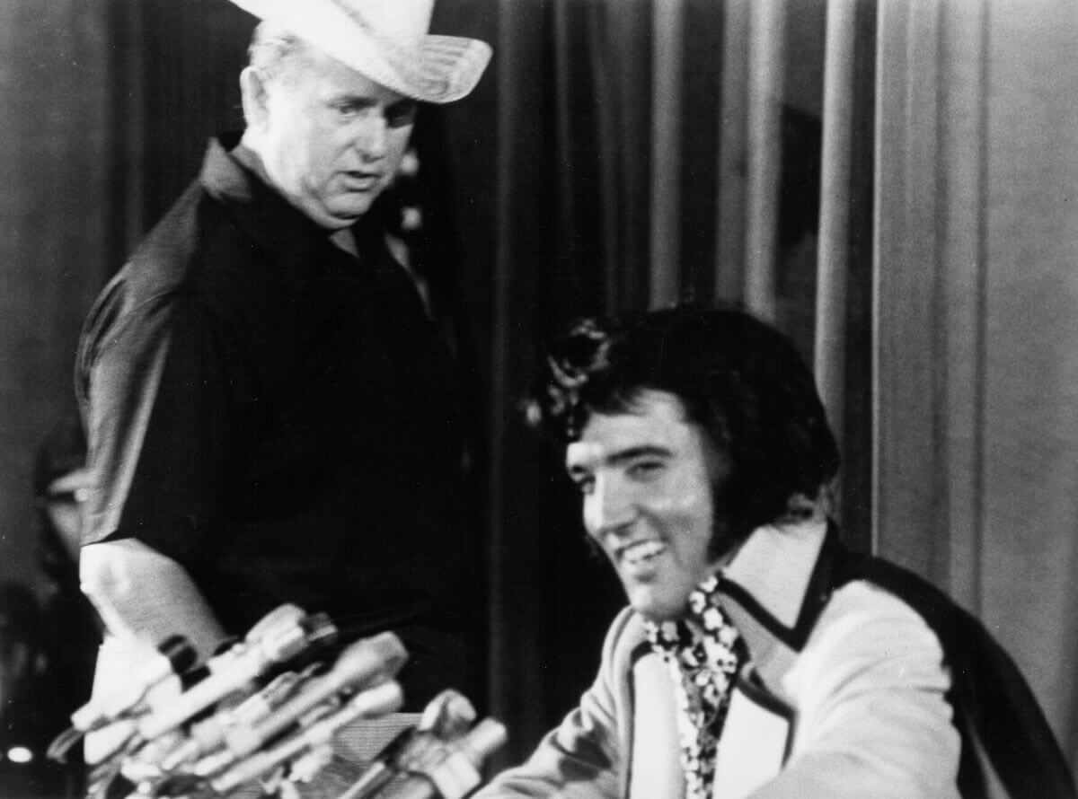 A black and white picture of Colonel Tom Parker wearing a cowboy hat and watching Elvis, who sits in front of multiple microphones.
