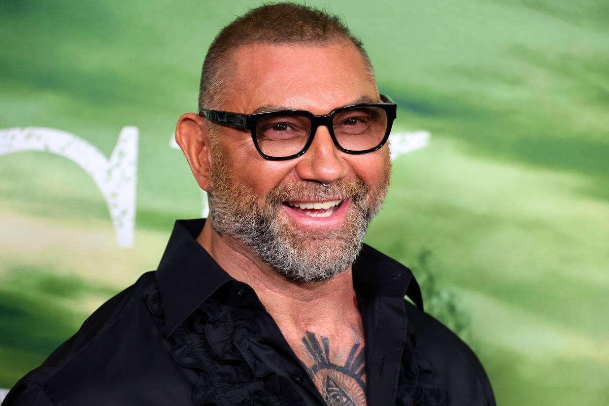 Dave Bautista wears glasses and smiles in front of a green background.