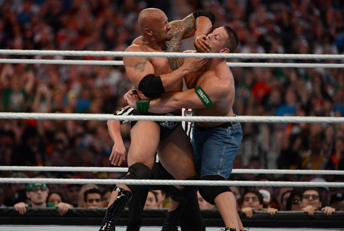 Dwayne Johnson and John Cena wrestle in the ring. Johnson has his hands on Cena's face and Cena has his arms wrapped around Johnson's waist.