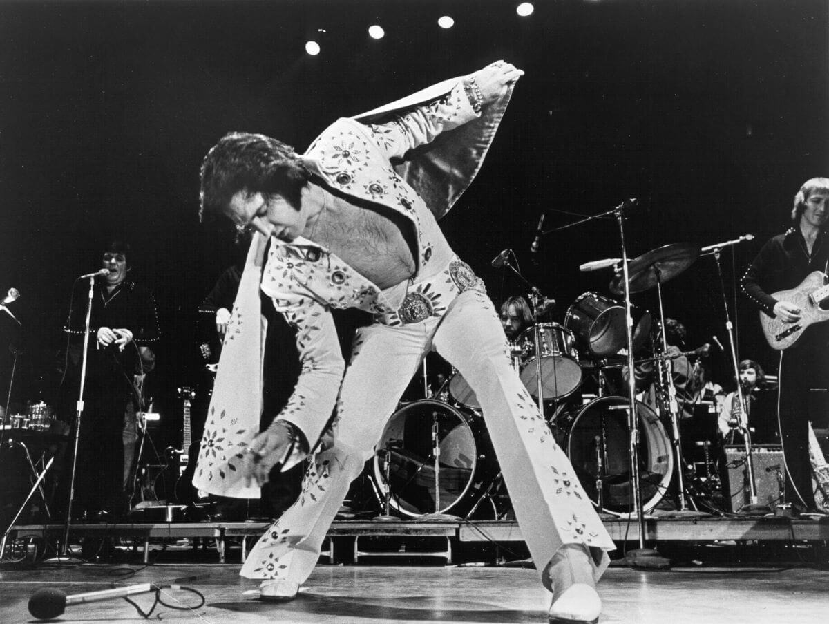 A black and white picture of Elvis wearing a jumpsuit and leaning forward while onstage.