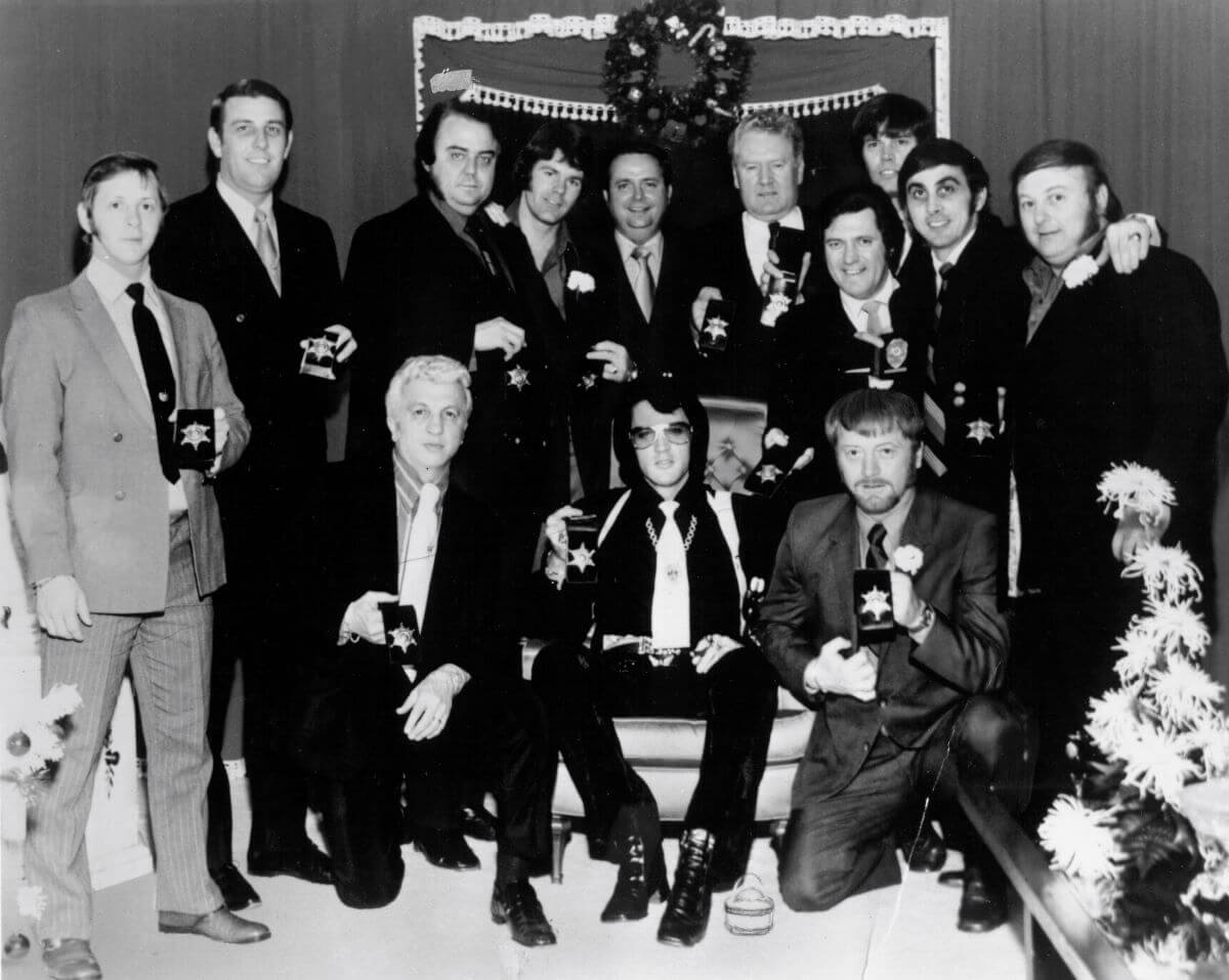 A black and white picture of Billy Smith, Bill Morris, Lamar Fike, Jerry Schilling, Sheriff Roy Nixon, Vernon Presley, Charlie Hodge, Sonny West, George Klein, Marty Lacker, Dr. George Nichopoulos, and Red West posing around Elvis Presley. Elvis sits in a chair.