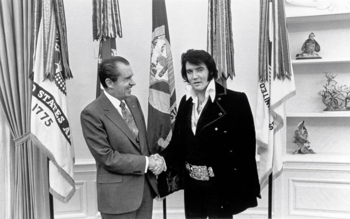 A black and white picture of Elvis and Richard Nixon shaking hands in front of flags.