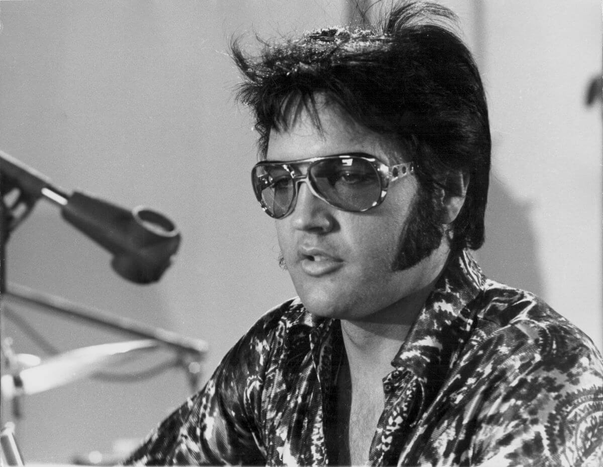 A black and white picture of Elvis sitting and wearing sunglasses.