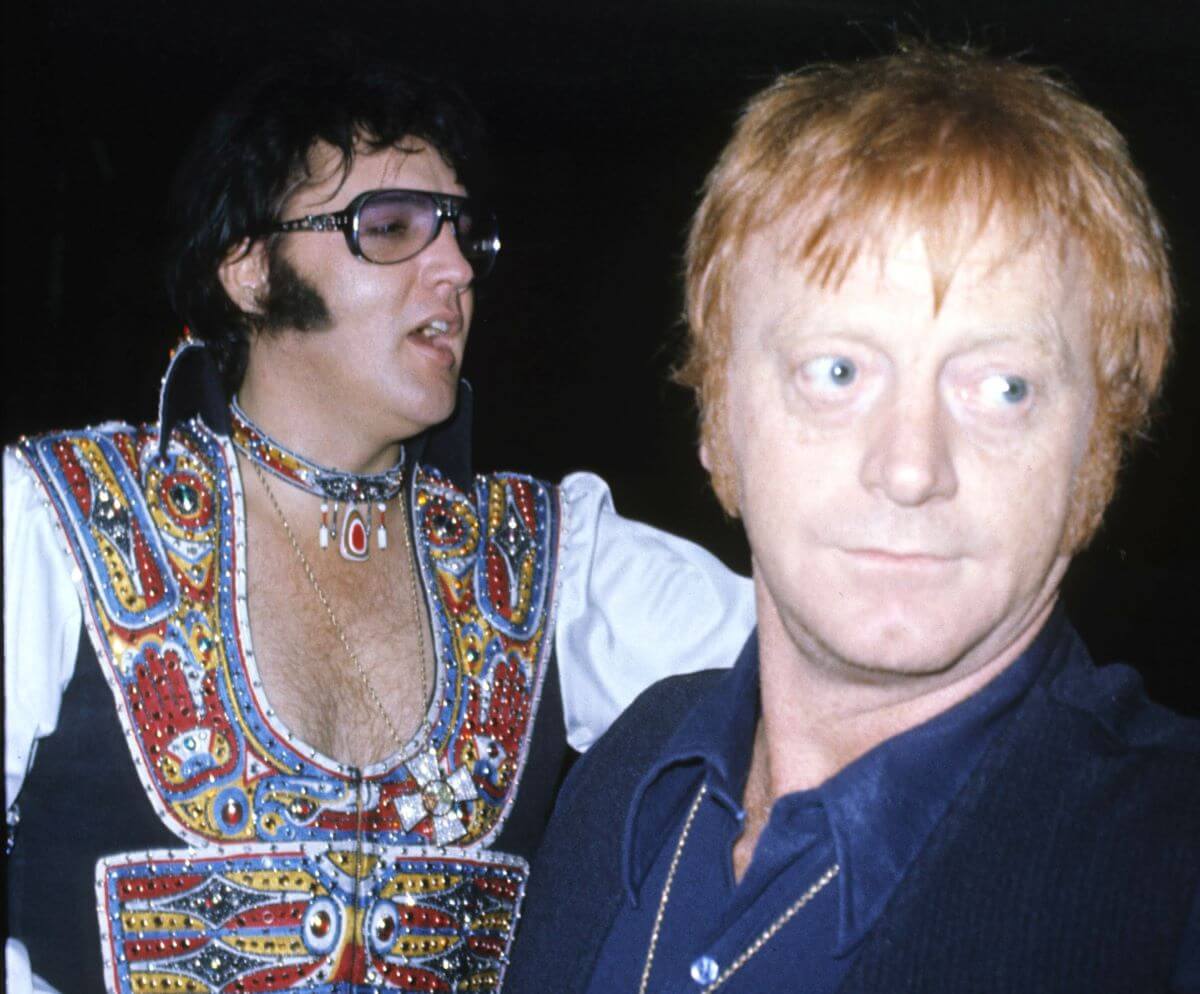 Elvis wears a multicolored blouse and a matching necklace and stands behind Red West, who wears a blue shirt.