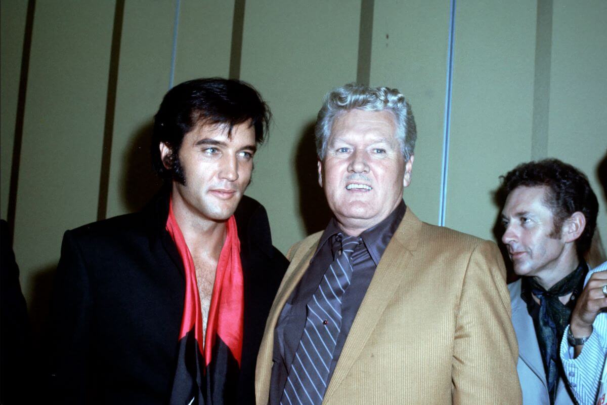 Elvis and Vernon Presley stand next to each other. Elvis wears a black suit and red scarf and Vernon wears a brown jacket and black shirt.