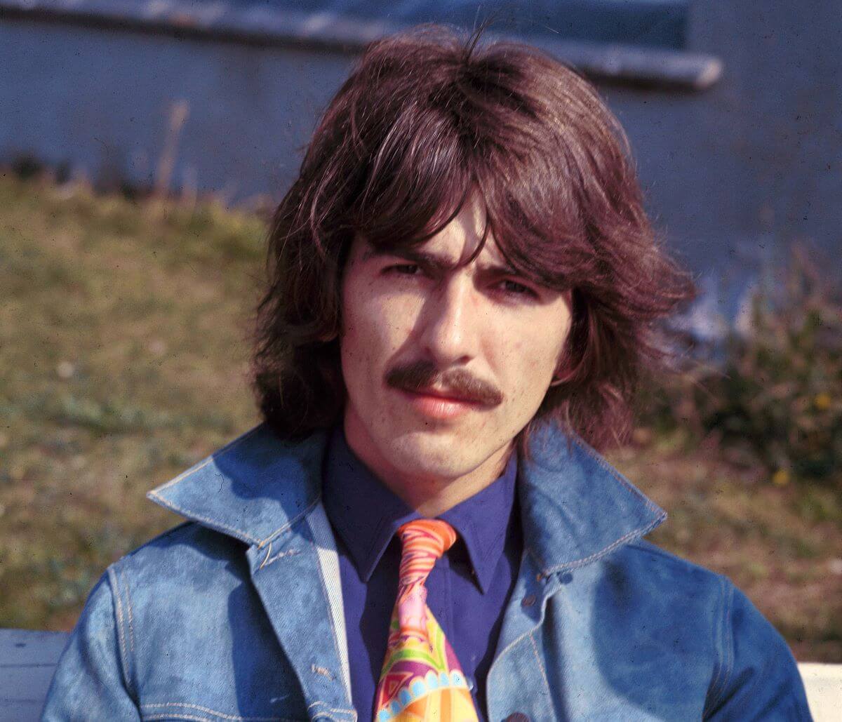The Beatles' George Harrison wears a denim jacket and a tie. He sits on the grass near water.