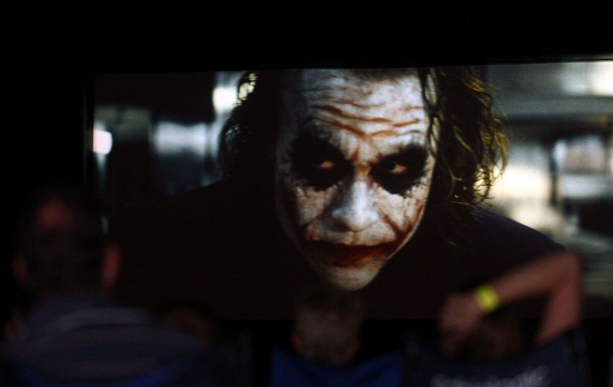 Heath Ledger in makeup as the Joker is on a screen while blurred figures look on.