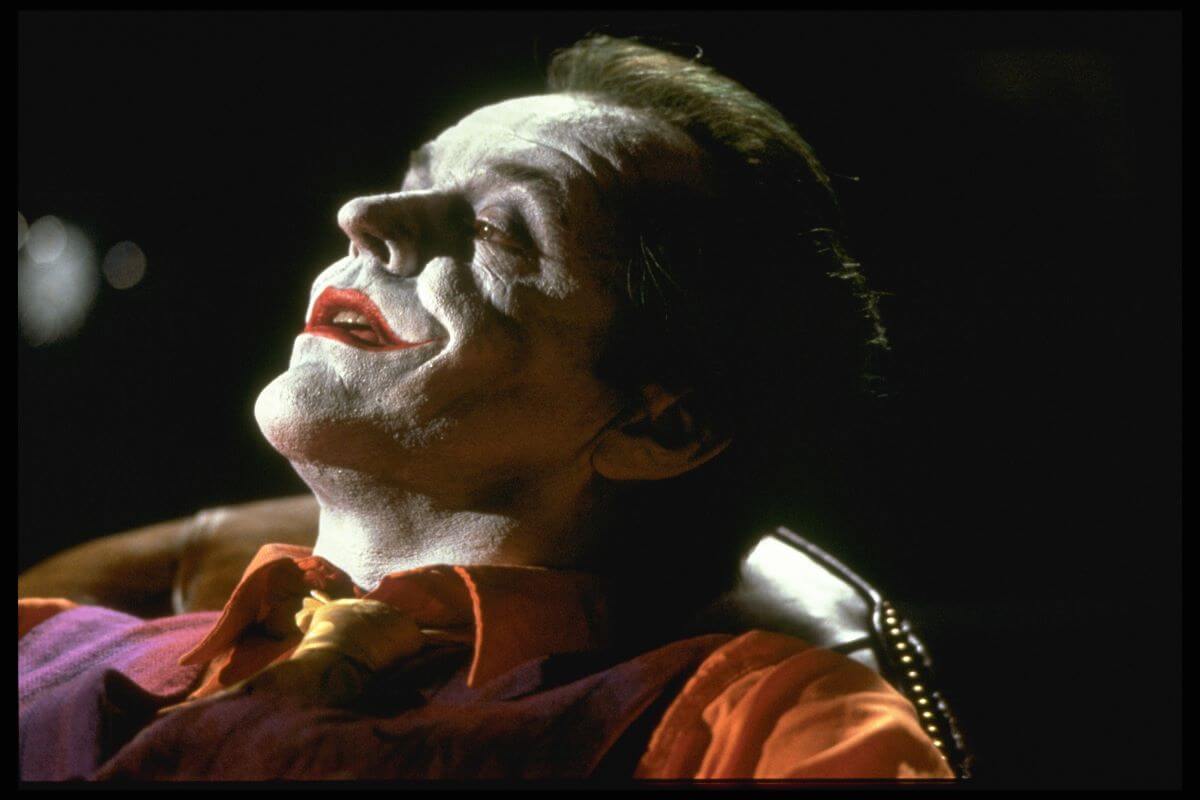 Jack Nicholson wears his Joker makeup and leans back in a chair.
