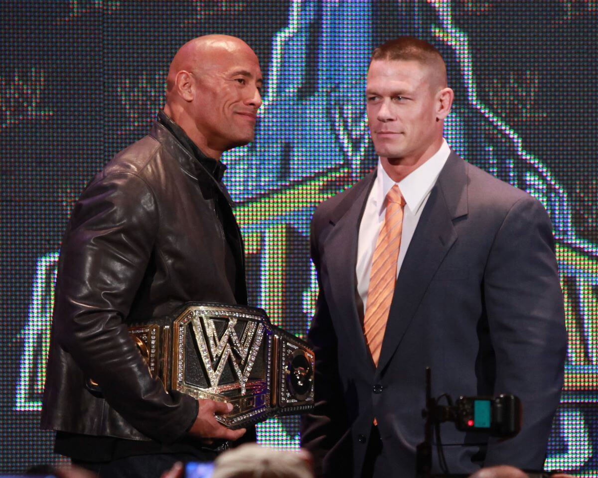 Dwayne Johnson holds a WWE title belt and stands next to John Cena, who wears a suit and tie.