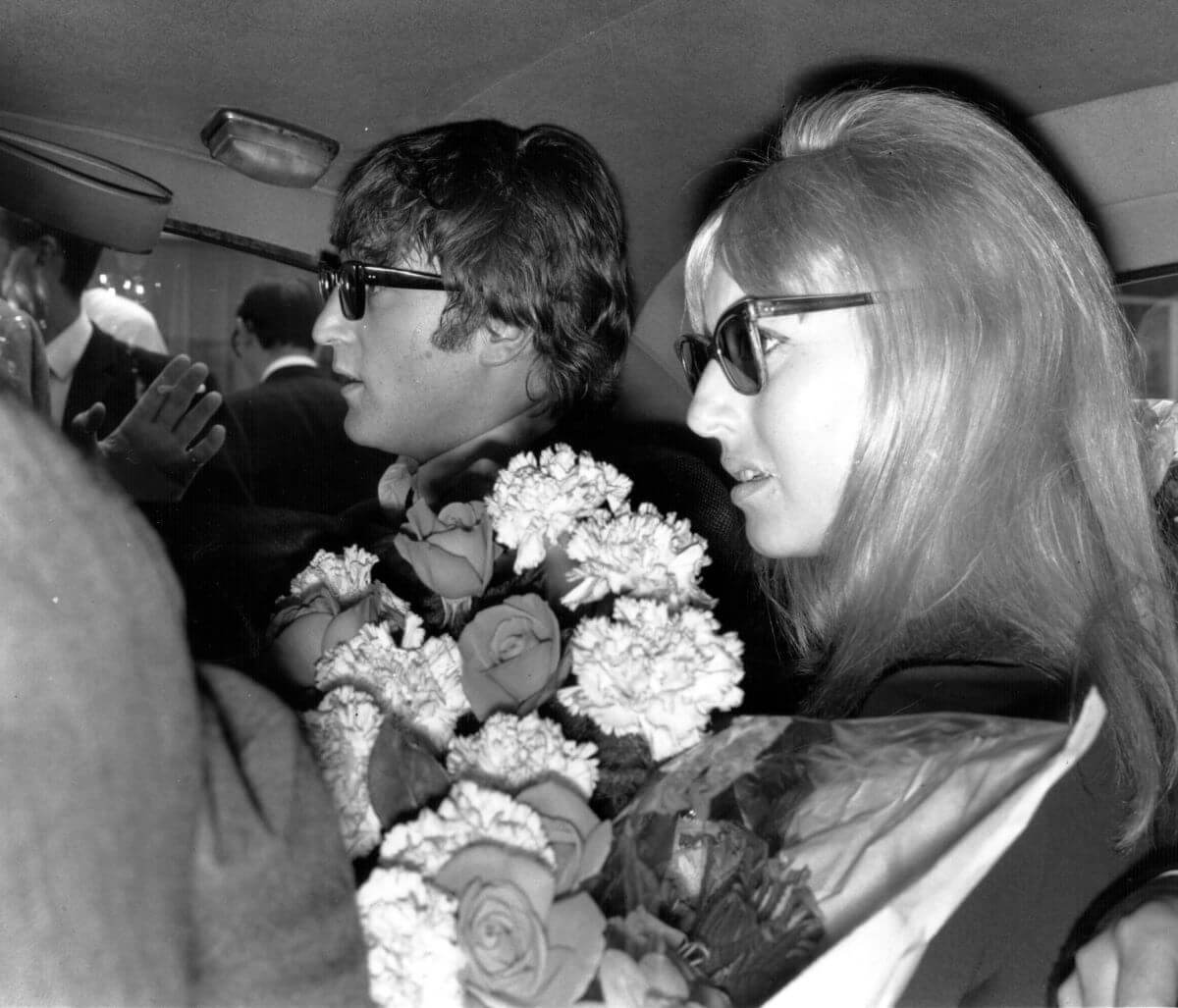 A black and white picture of John and Cynthia Lennon wearing sunglasses in the backseat of a car. Cynthia holds a bouquet of flowers.