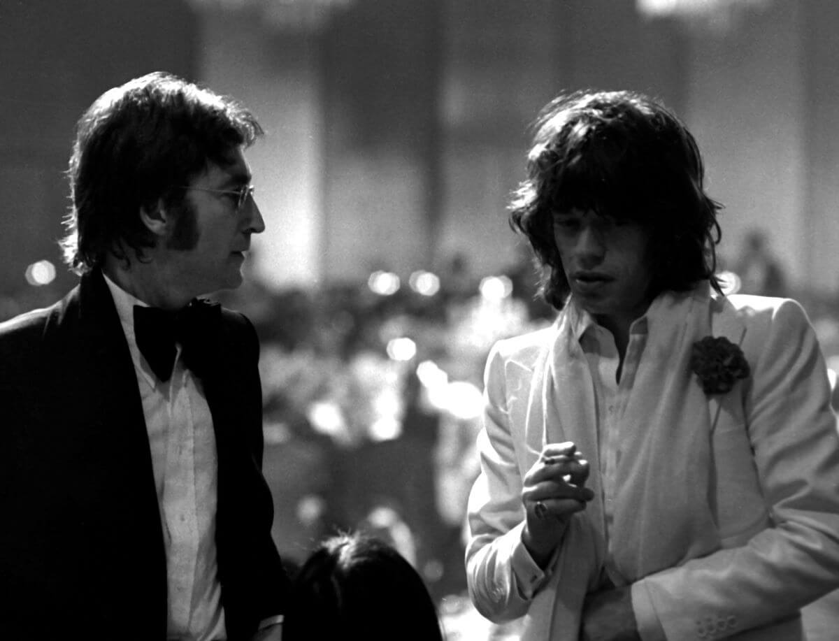 A black and white picture of John Lennon wearing a tuxedo and talking to Mick Jagger, who wears a white suit and holds a cigarette.