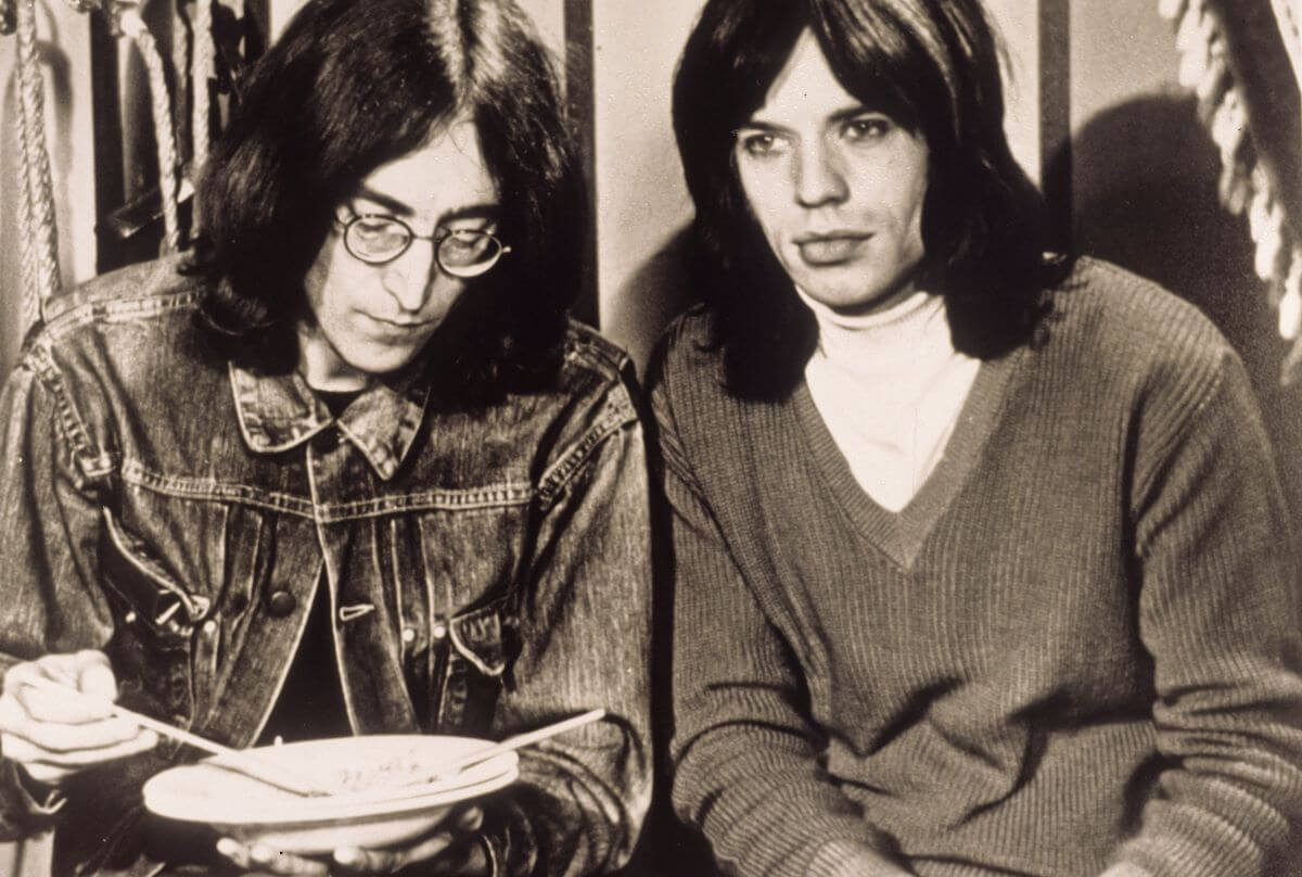 John Lennon holds a plate and sits next to The Rolling Stones' Mick Jagger.