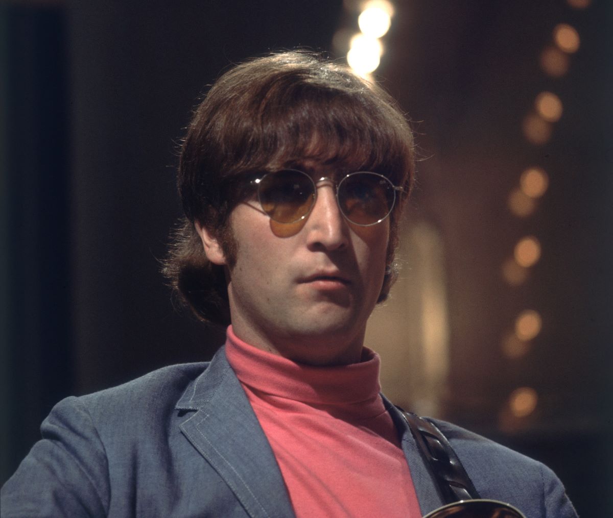 John Lennon wears a red turtleneck, blue jacket, and sunglasses.