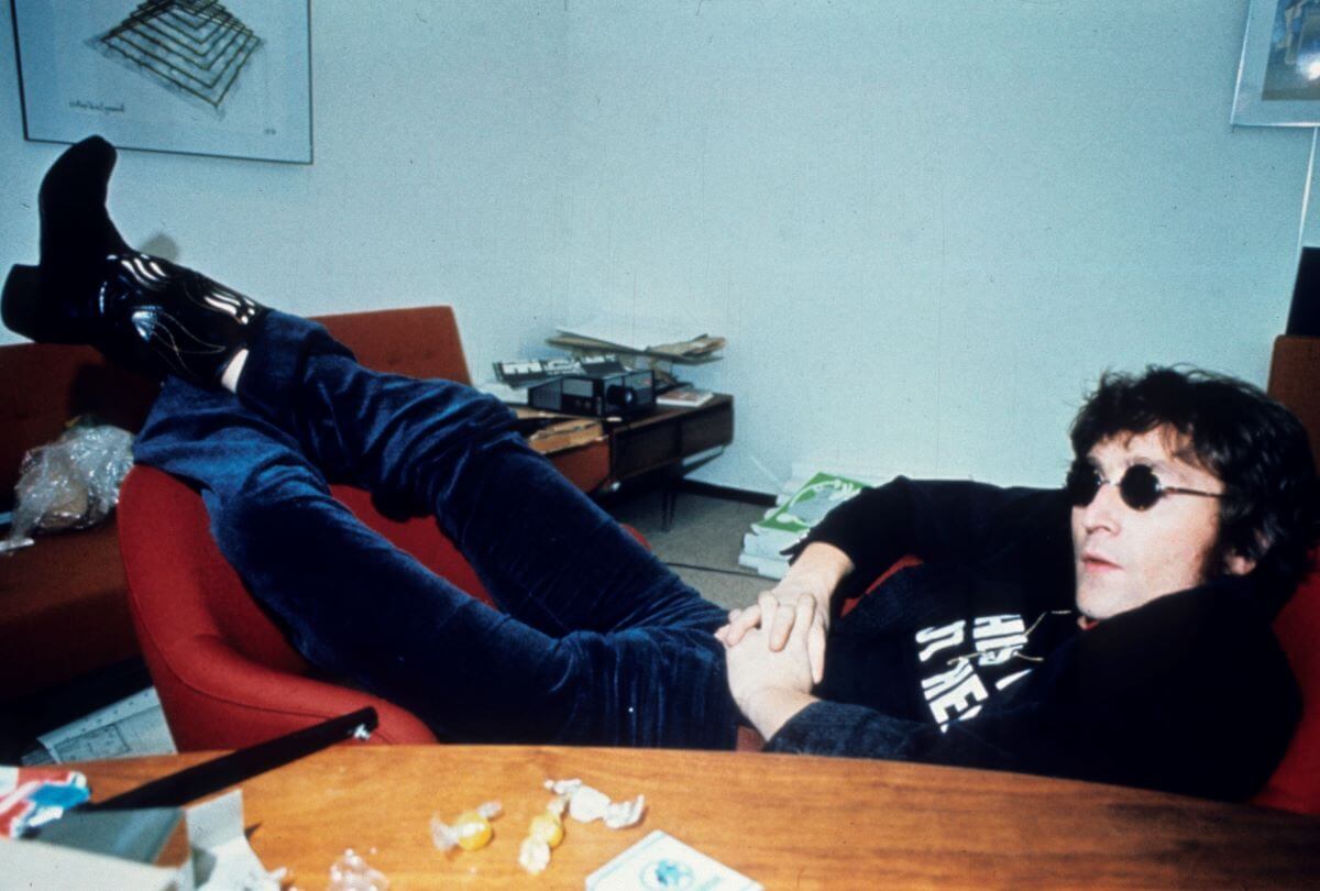 John Lennon slouches in one red chair and leans his legs on another chair. He wears sunglasses and sits in front of a table with hard candy.