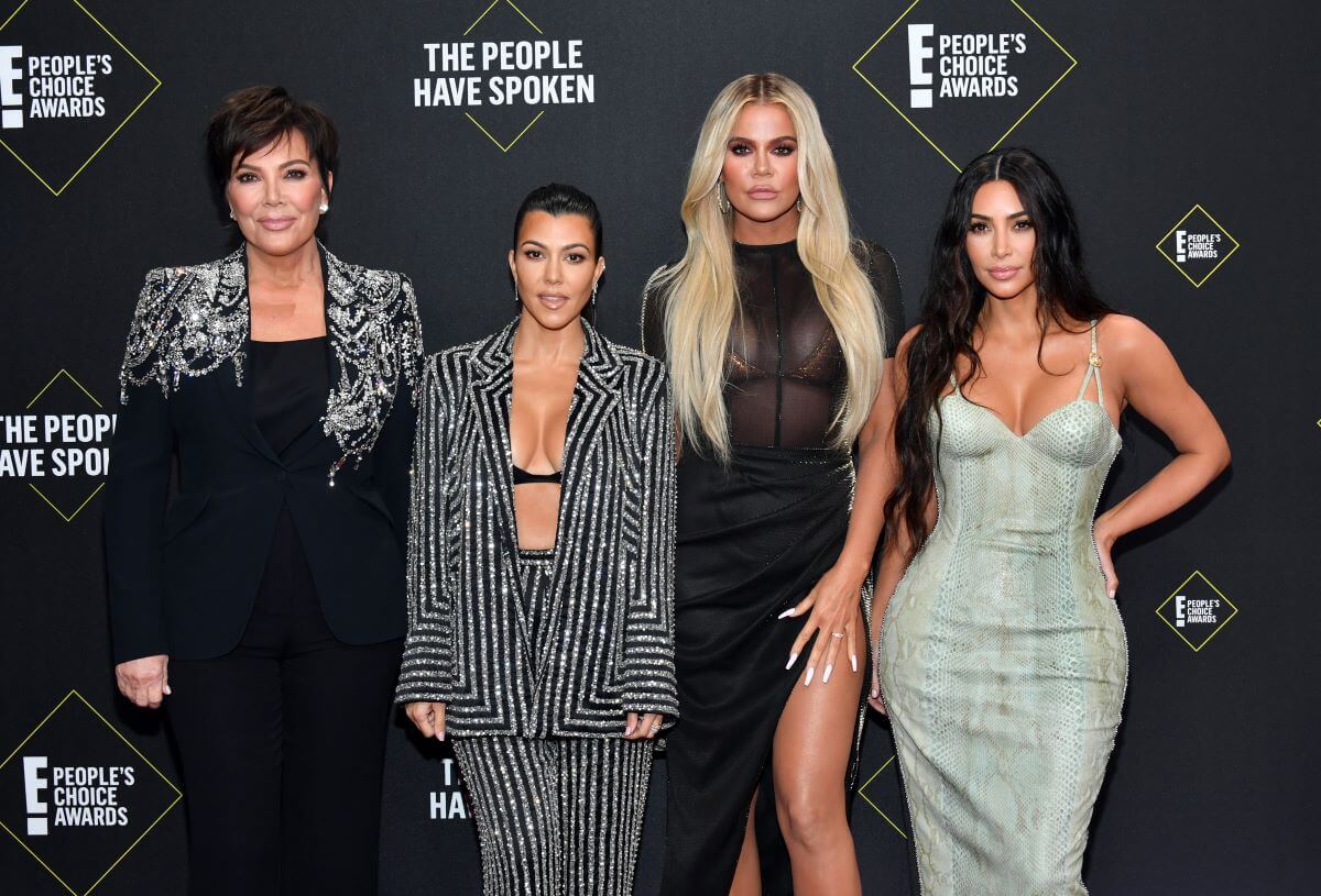 Kris Jenner, Kourtney Kardashian, Khloé Kardashian, and Kim Kardashian stand in a line in front of signage for the E! People's Choice Awards.