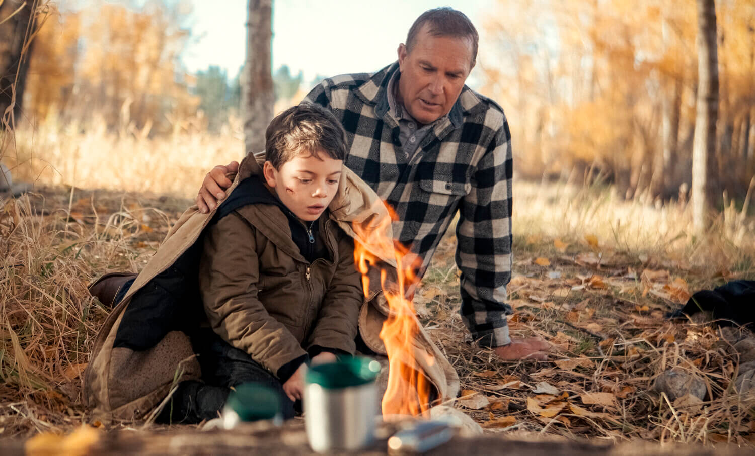 Brecken Merrill as Tate Dutton and Kevin Costner as John Dutton sitting next to a fire outside in 'Yellowstone'