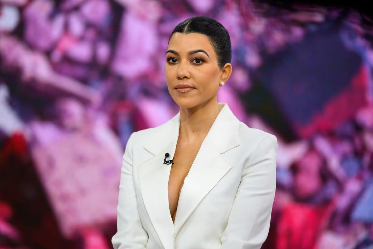 Kourtney Kardashian wears a white blazer and sits in front of a purple background.
