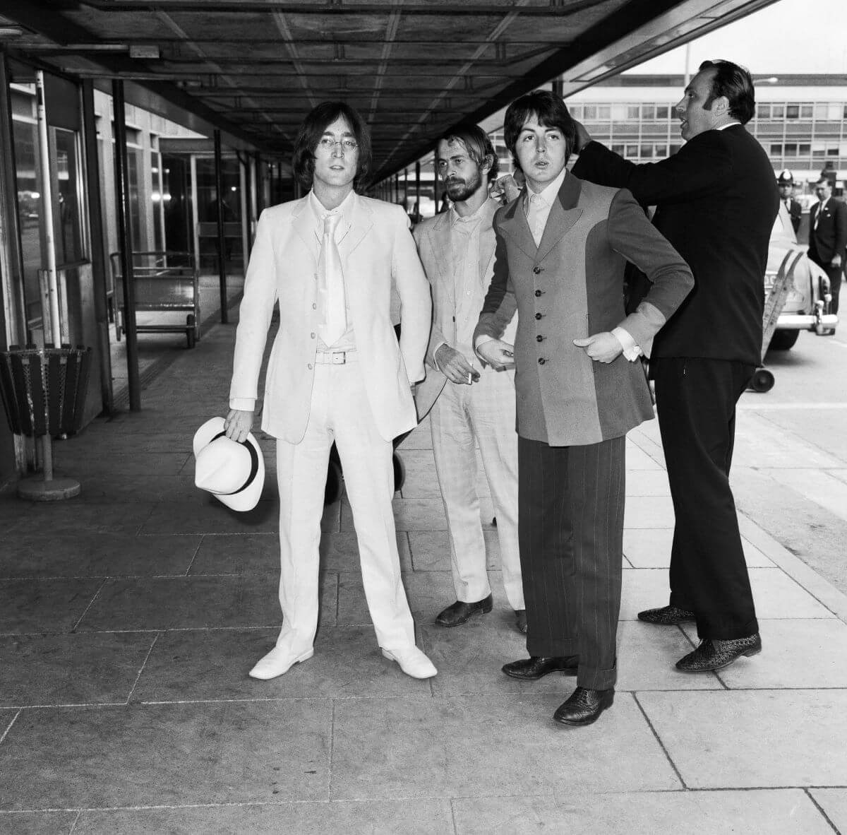A black and white picture of John Lennon, Alex Mardas, Paul McCartney, and Les Anthony standing outside an airport. Lennon holds a hat.