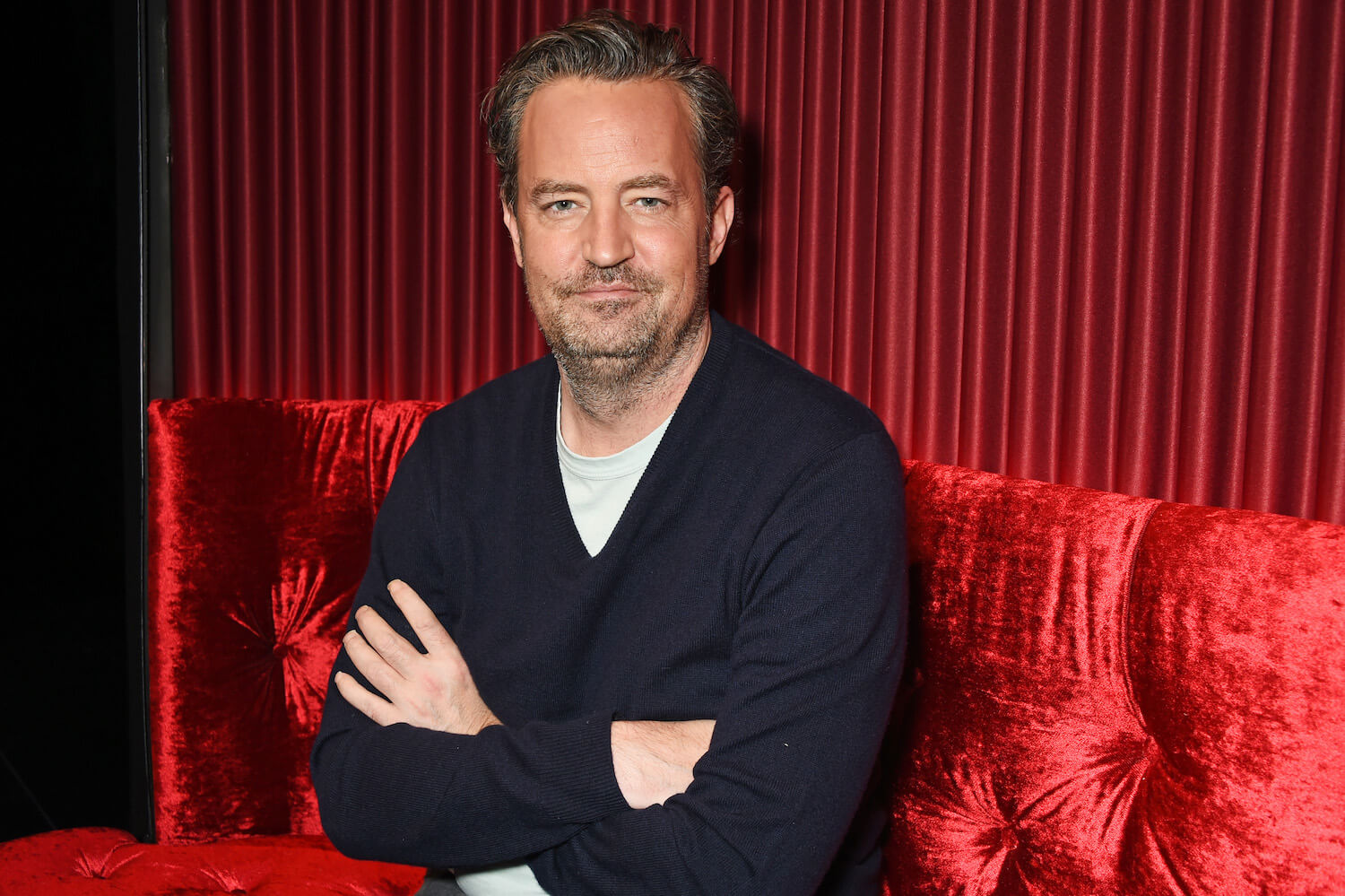 'Friends' star Matthew Perry sitting in front of a red curtain with his arms crossed
