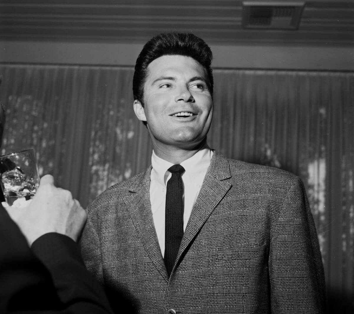 A black and white picture of Max Baer Jr. of 'The Beverly Hillbillies' wearing a suit as someone takes his picture.
