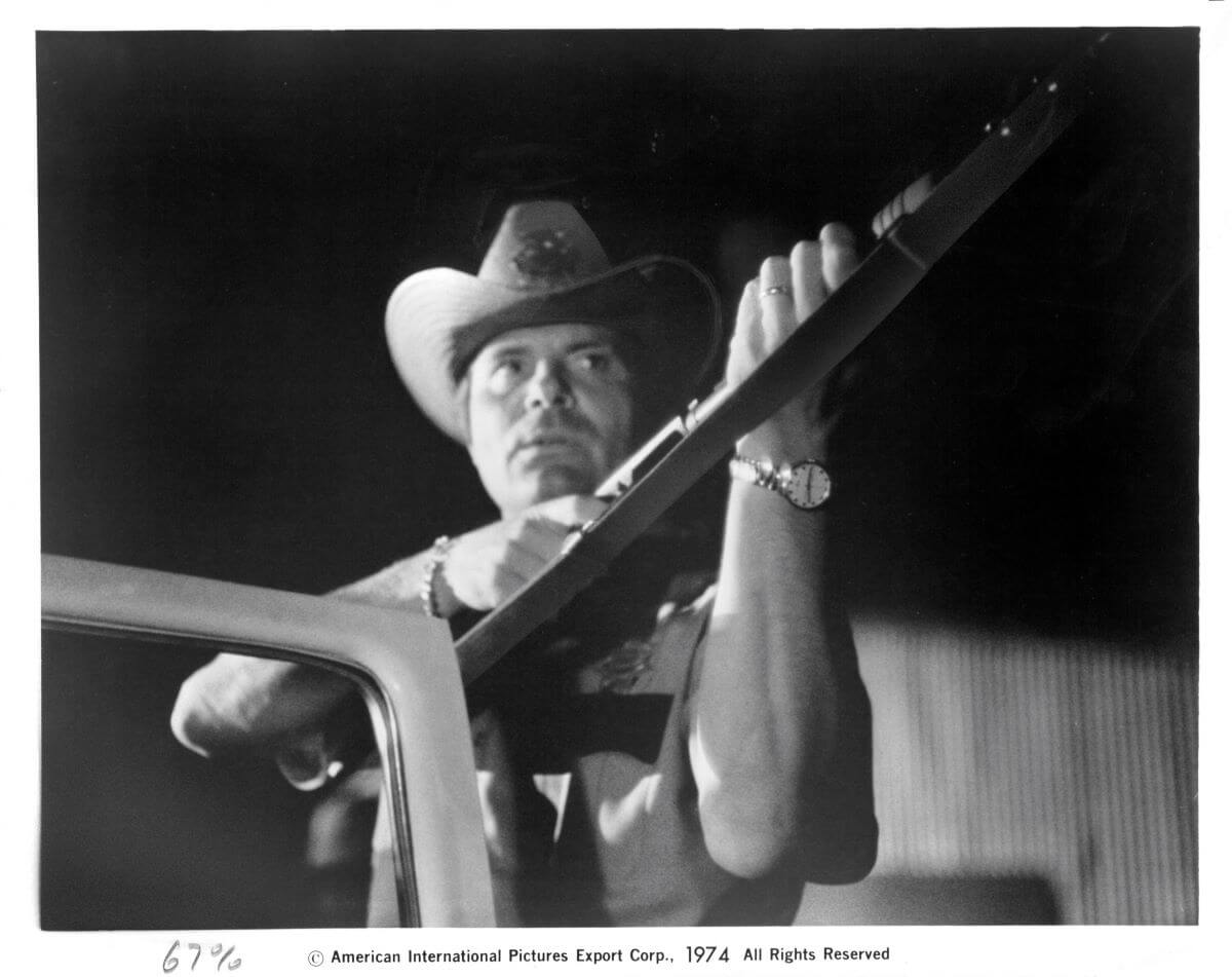 A black and white picture of Max Baer Jr. holding a gun while wearing a sheriff's uniform in 'Macon County Line.'