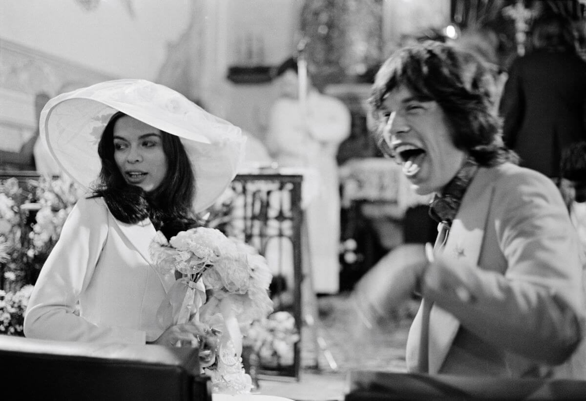 A black and white picture of Bianca Jagger holding a bouquet and sitting at a church pew next to Mick Jagger, who laughs.