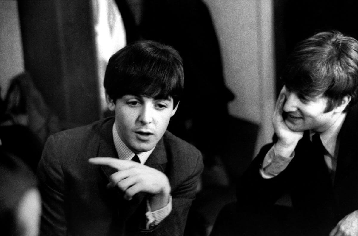A black and white picture of Paul McCartney talking while John Lennon rests his head in his hand and watches him.