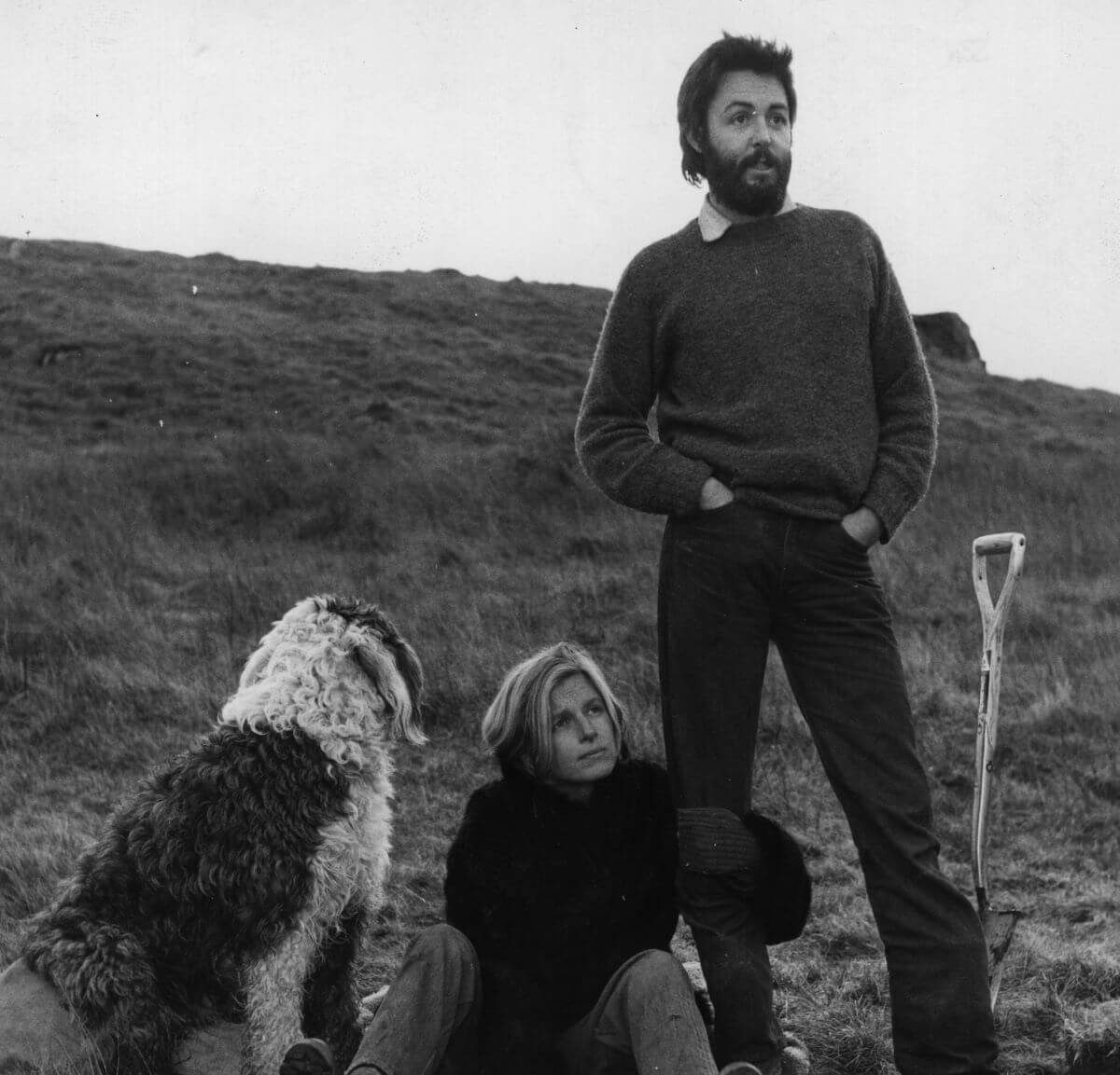A black and whtie picture of Paul McCartney standing next to a shovel. Linda McCartney sits at his feet next to their dog.