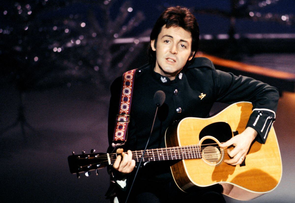 Paul McCartney strums an acoustic guitar and stands in front of a microphone.