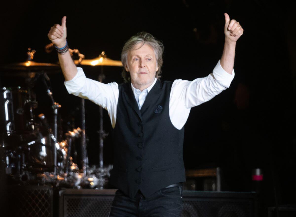 Paul  McCartney stands onstage and lifts his arms up to give two thumbs up.