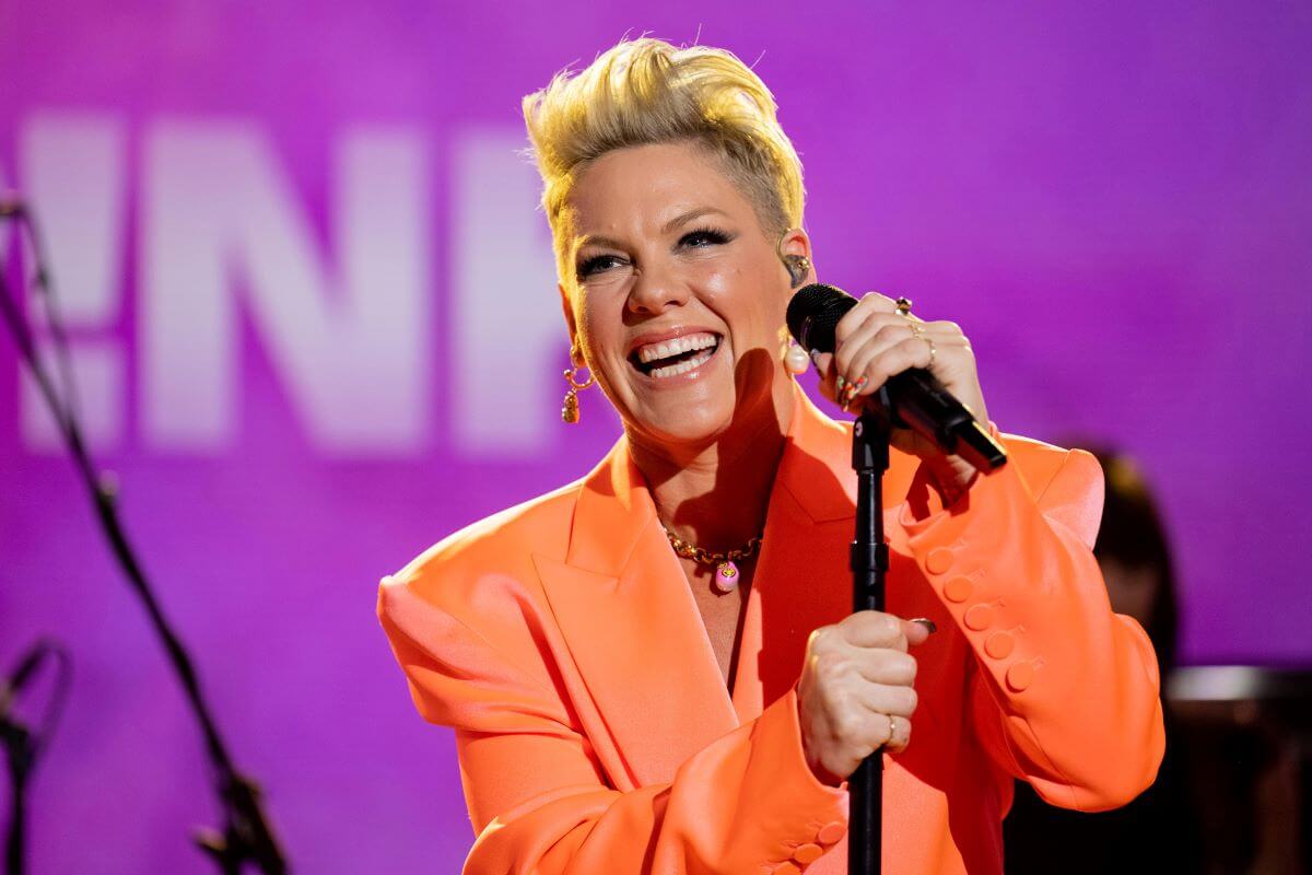 Pink wears an orange blazer and holds a microphone.