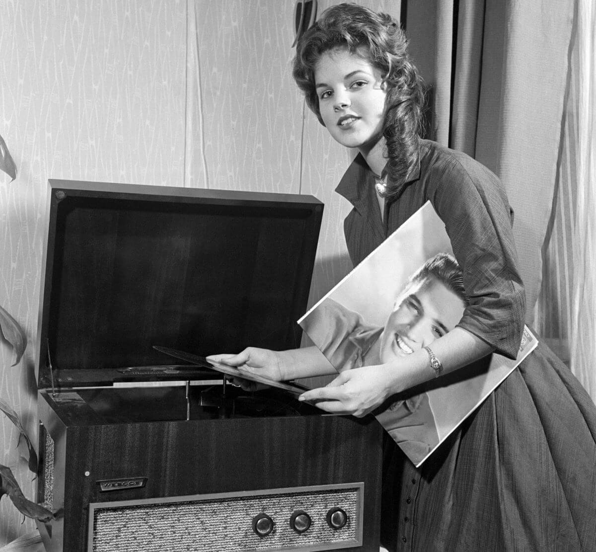 A black and white picture of Priscilla standing over a record player and holding an Elvis record under her arm.