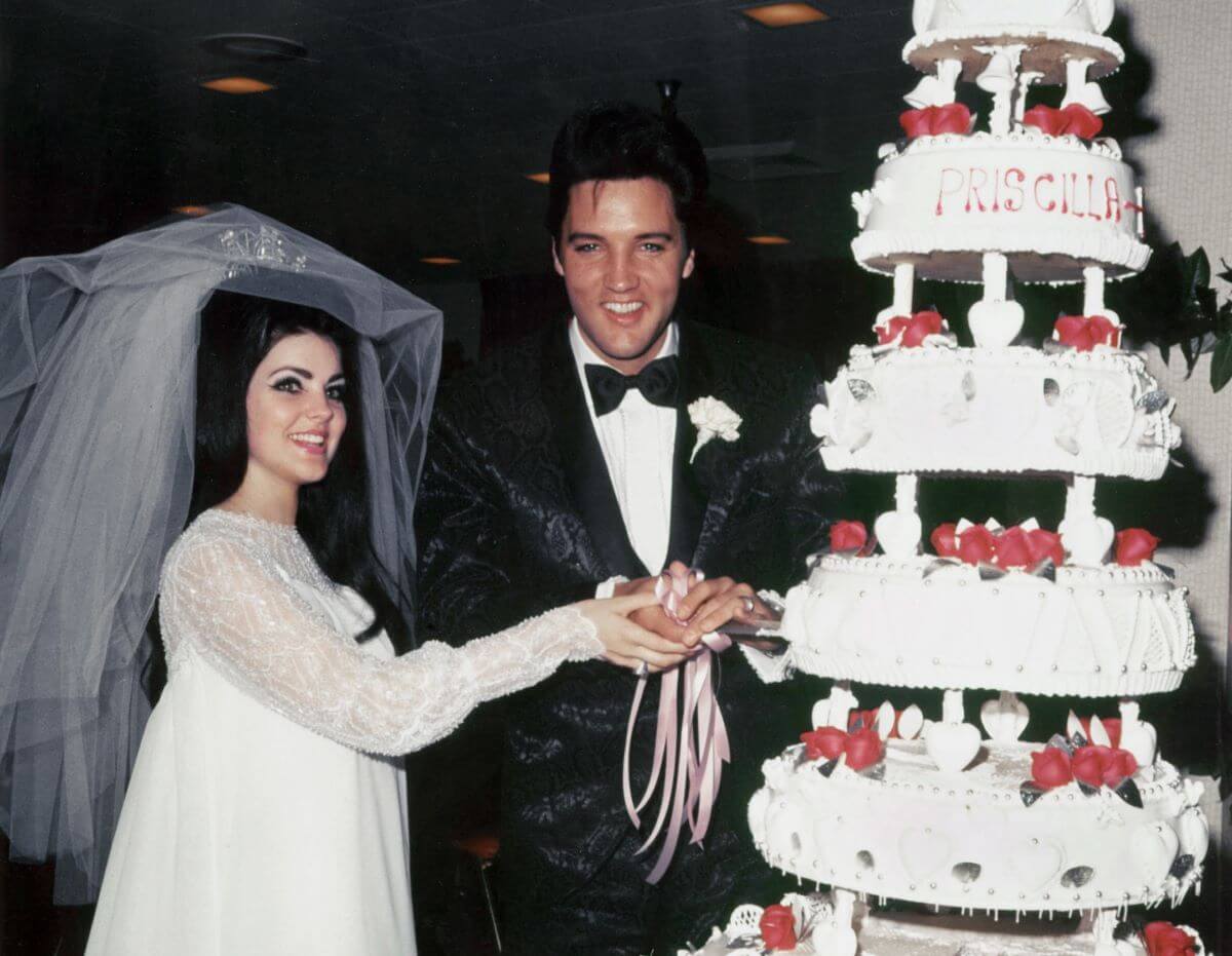 Priscilla and Elvis Presley cut into their wedding cake. She wears a dress and veil and he wears a tuxedo.