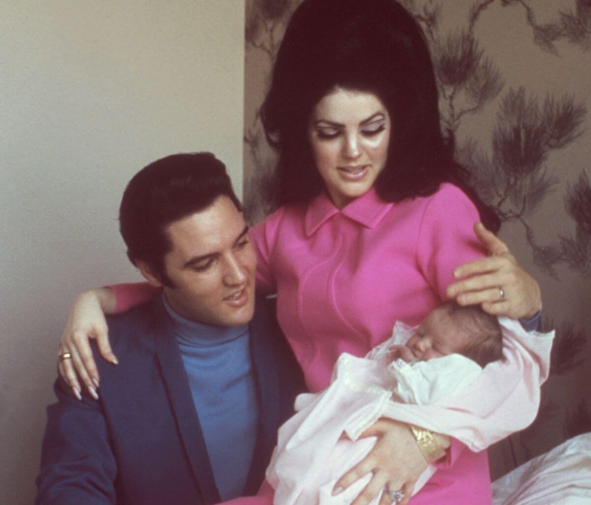 Priscilla Presley holds an infant Lisa Marie Presley while sitting on Elvis' lap. They both look at Lisa Marie. 