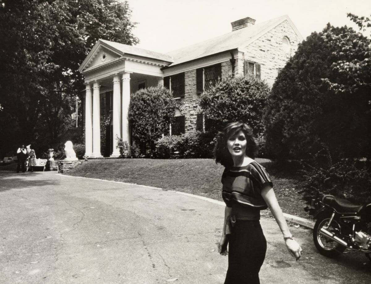 A black and white picture of Priscilla Presley walking up the driveway to Graceland, the house she shared with Elvis.