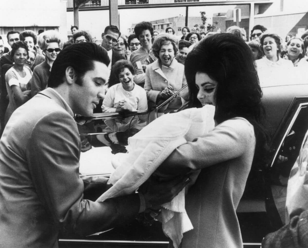 A black and white picture of Priscilla Presley holding Lisa Marie Presley. Elvis looks down at his daughter. The couple stands in front of fans.