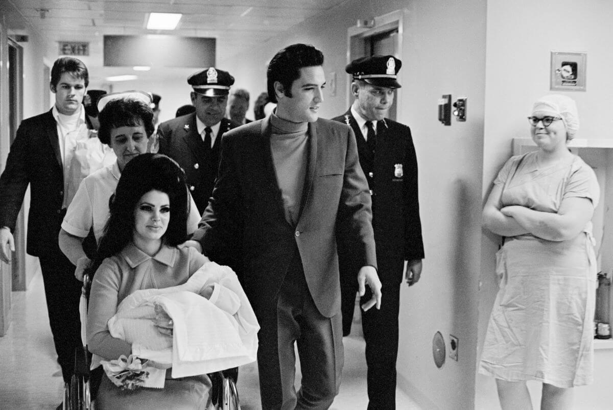A black and white picture of Priscilla Presley holding Lisa Marie Presley in her arms. A nurse pushes her in a wheelchair. She's accompanied by Elvis and a police escort.