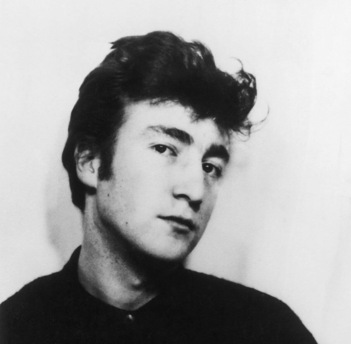 A black and white picture of John Lennon looking at the camera and holding his head at an angle.
