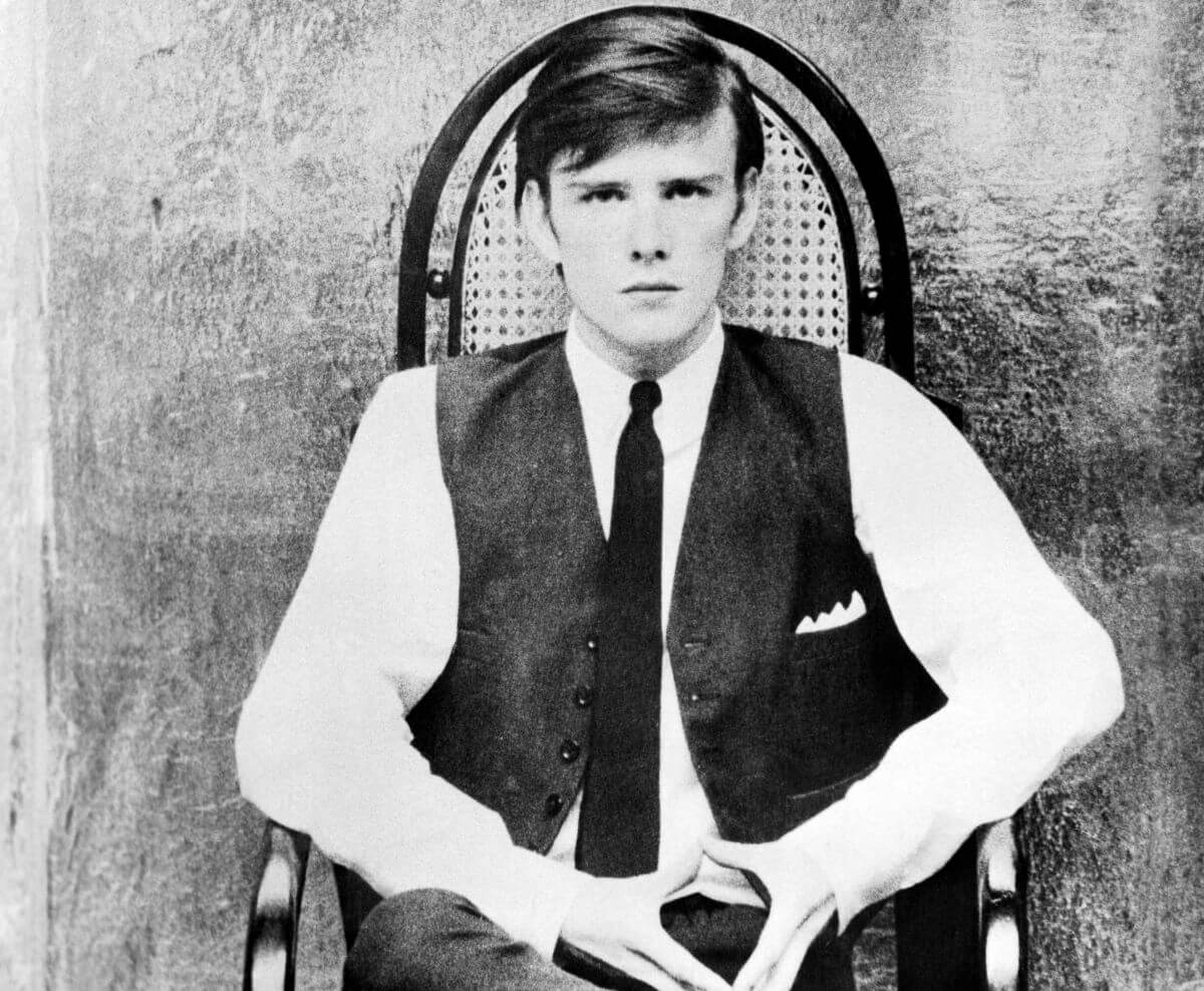 A black and white picture of Stuart Sutcliffe sitting in a chair and wearing a tie and vest.