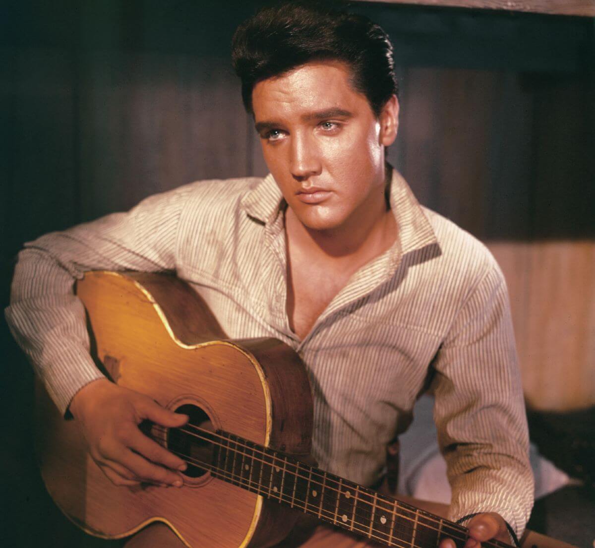 Elvis Presley sits wearing a buttoned shirt and holds an acoustic guitar.