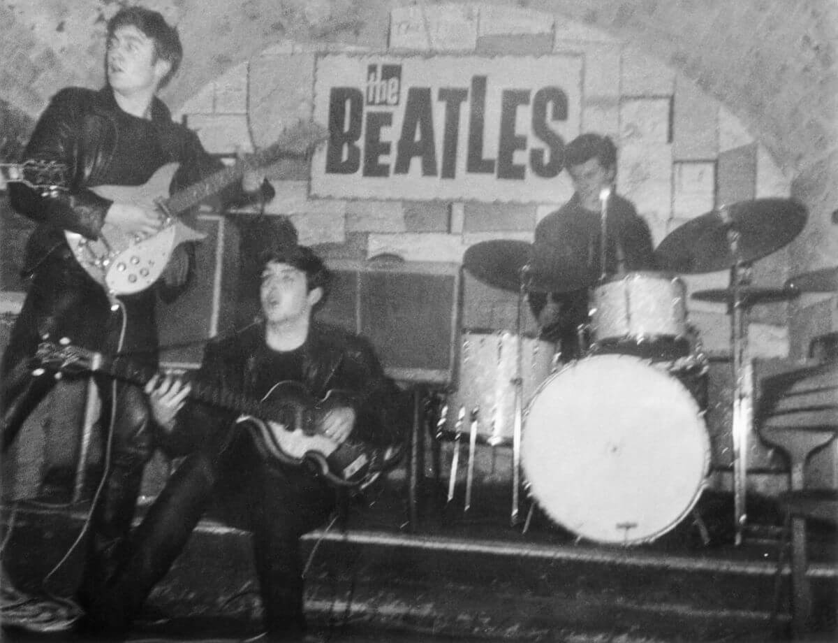 A black and white picture of John Lennon, Paul McCartney, and drummer Pete Best performing in front of a sign for "The Beatles." Lennon and McCartney both play guitars and McCartney sits down.