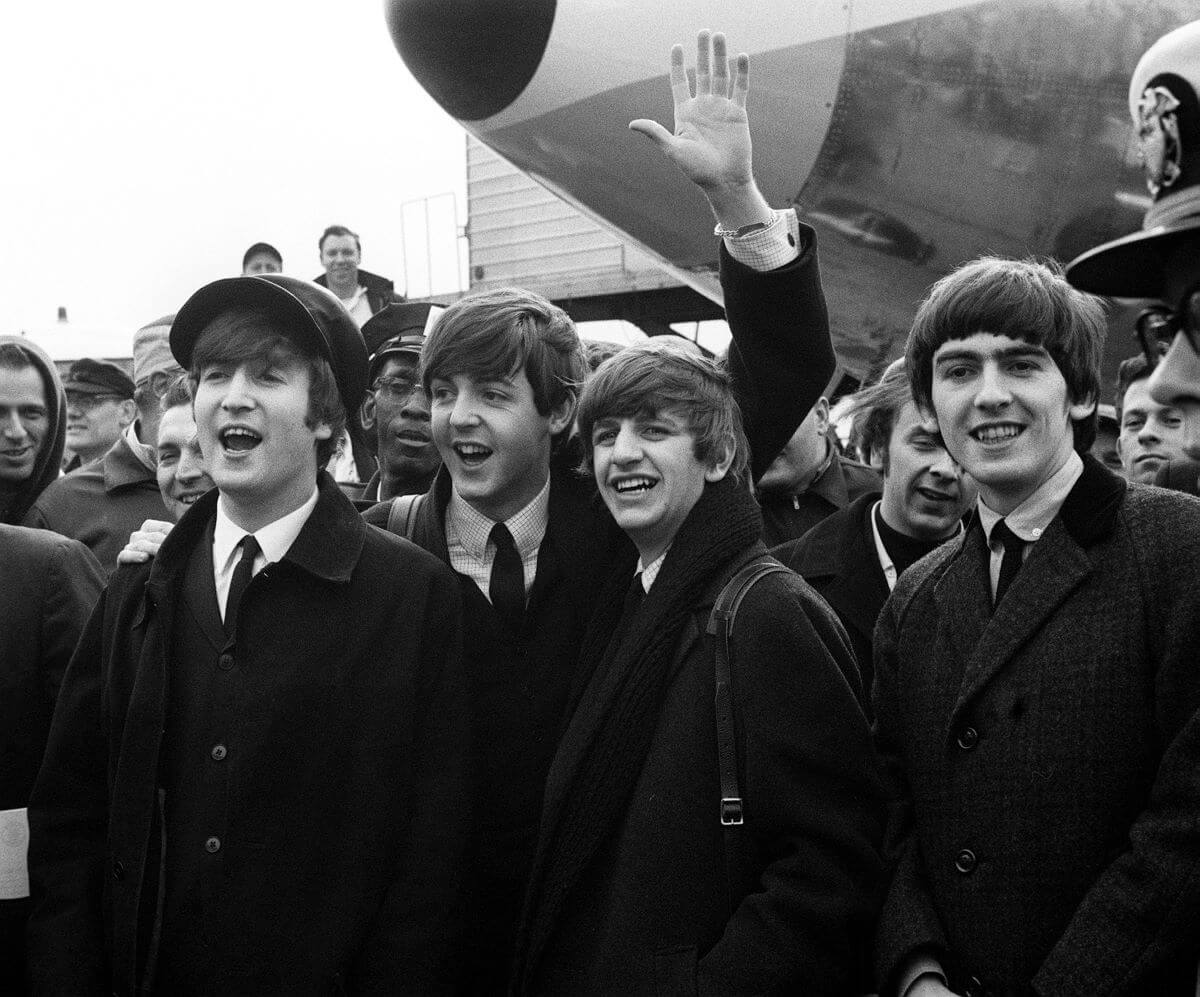 A black and white picture of Beatles band members John Lennon, Paul McCartney, Ringo Starr, and George Harrison standing at a crowded airport.