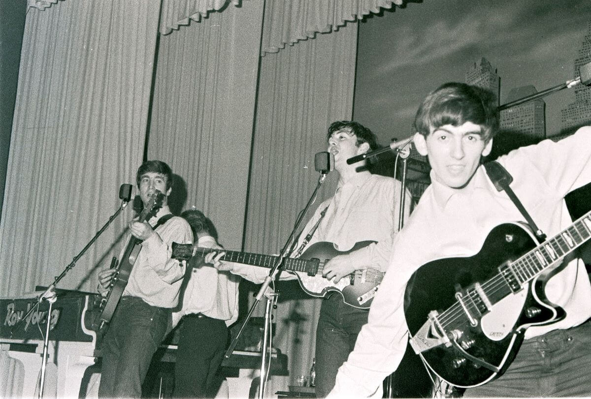 A black and white picture of John Lennon, Paul McCartney, and George Harrison on stage. Harrison spreads his arms wide and looks at the camera.