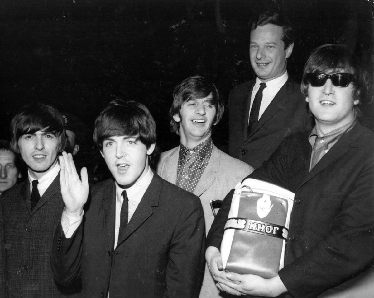 A black and white picture of George Harrison, Paul McCartney, Ringo Starr, Brian Epstein, and John Lennon. McCartney waves and Lennon carries a bag and wears sunglasses.