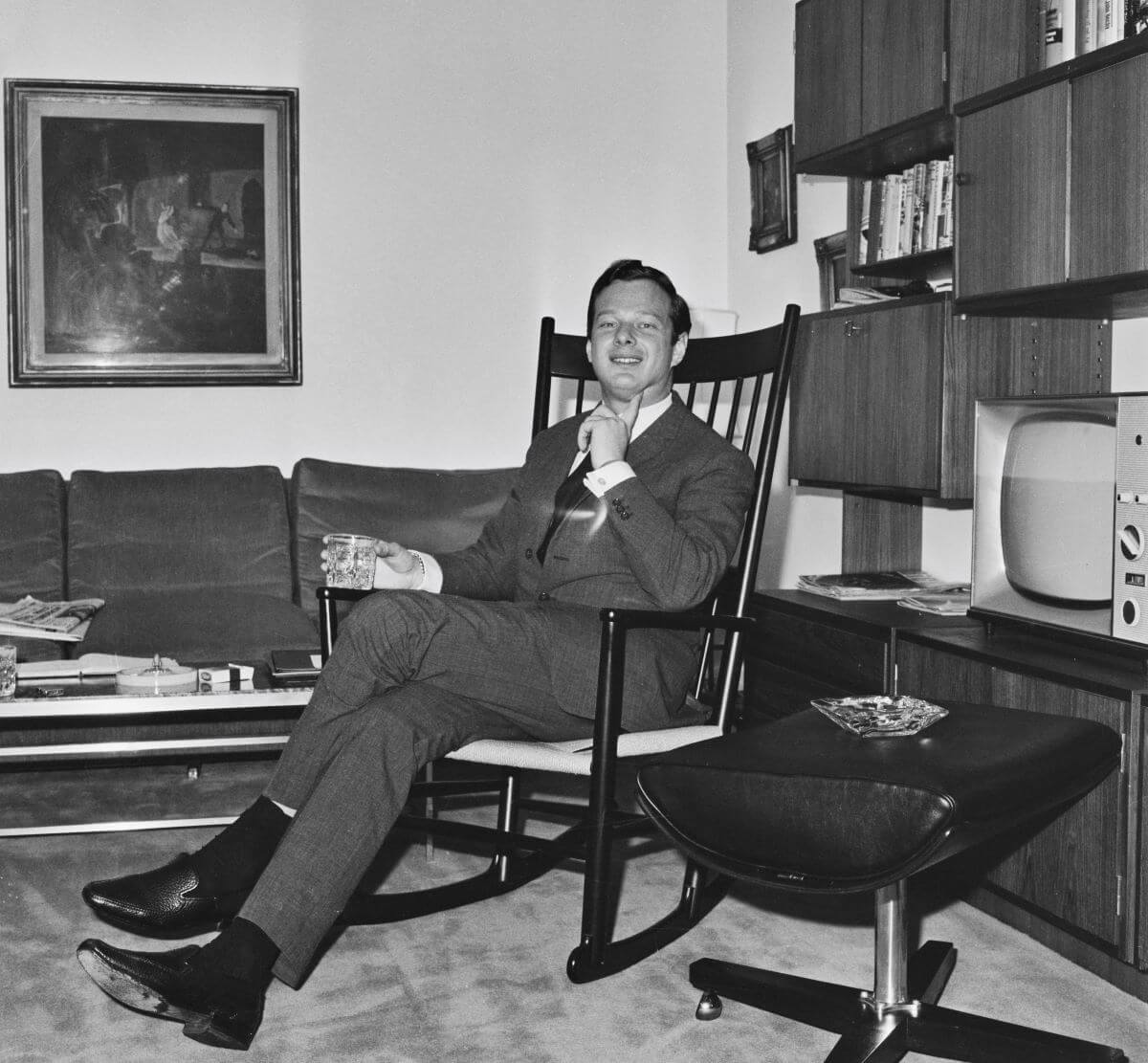 A black and white picture of Brian Epstein wearing a suit and sitting in a rocking chair. He holds a glass.