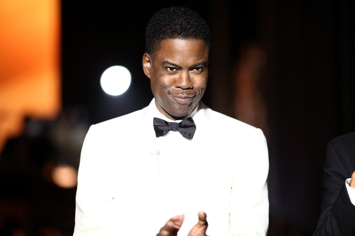 Chris Rock clapping on stage while hosting the 88th Academy Awards in a white suit.